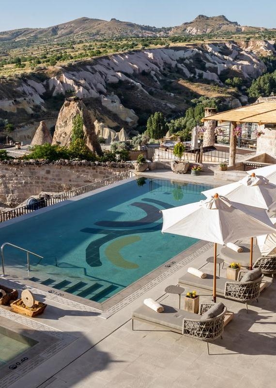 A blue infinity pool with lounge chairs and umbrellas overlooking the rugged Cappadocia landscape.