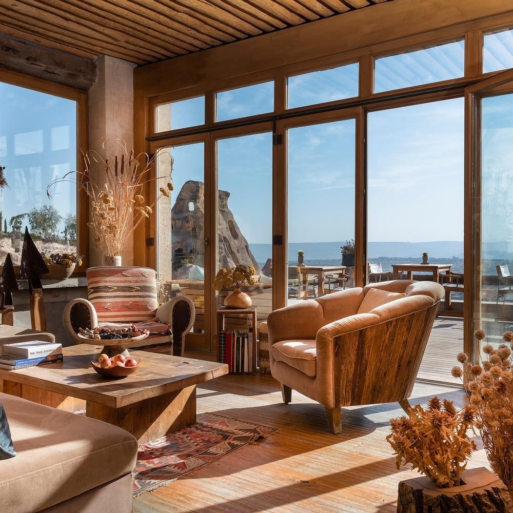 Sunlit lounge with floor-to-ceiling windows, wooden furniture, and a view of rock formations.