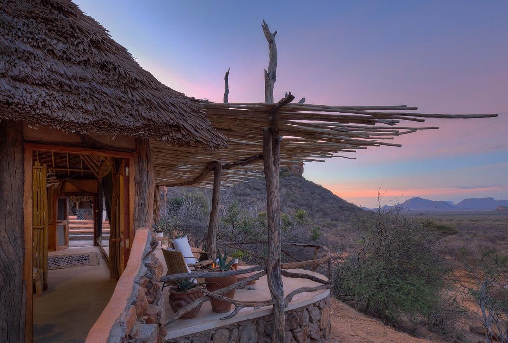 A balcony with a thatched roof and wooden branch pergola overlooking a valley at purple twilight.