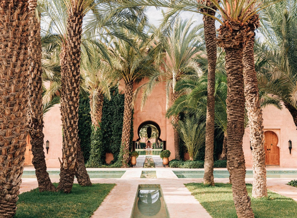 Tall palm trees and pink-walled buildings surrounding a narrow water feature in a tranquil Moroccan courtyard.