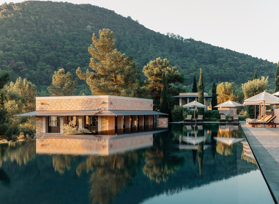 A stone pavilion at Amanruya Bodrum reflected in a still infinity pool, surrounded by pine trees and hills at dusk.