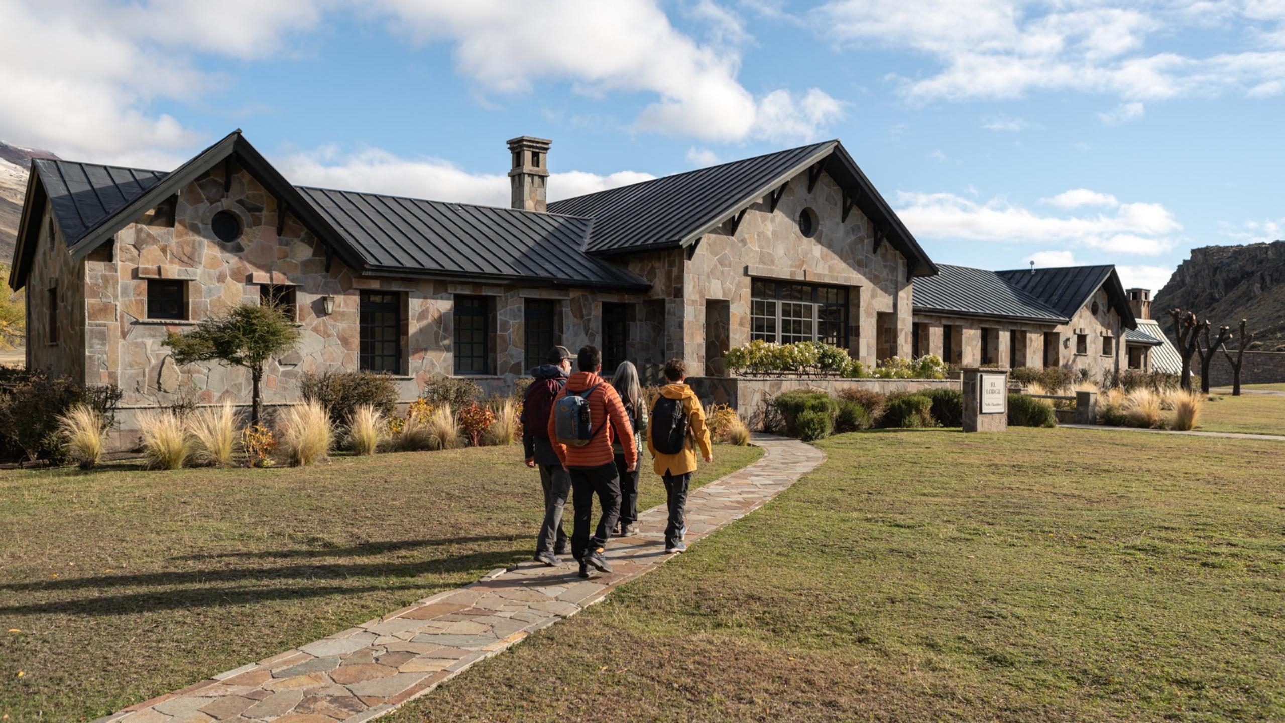 Four hikers with backpacks walking toward a stone lodge building at Explora Patagonia National Park.