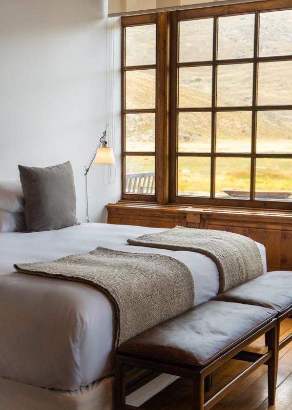 A bed with neutral linens and a leather bench next to a large window at Explora Patagonia National Park.