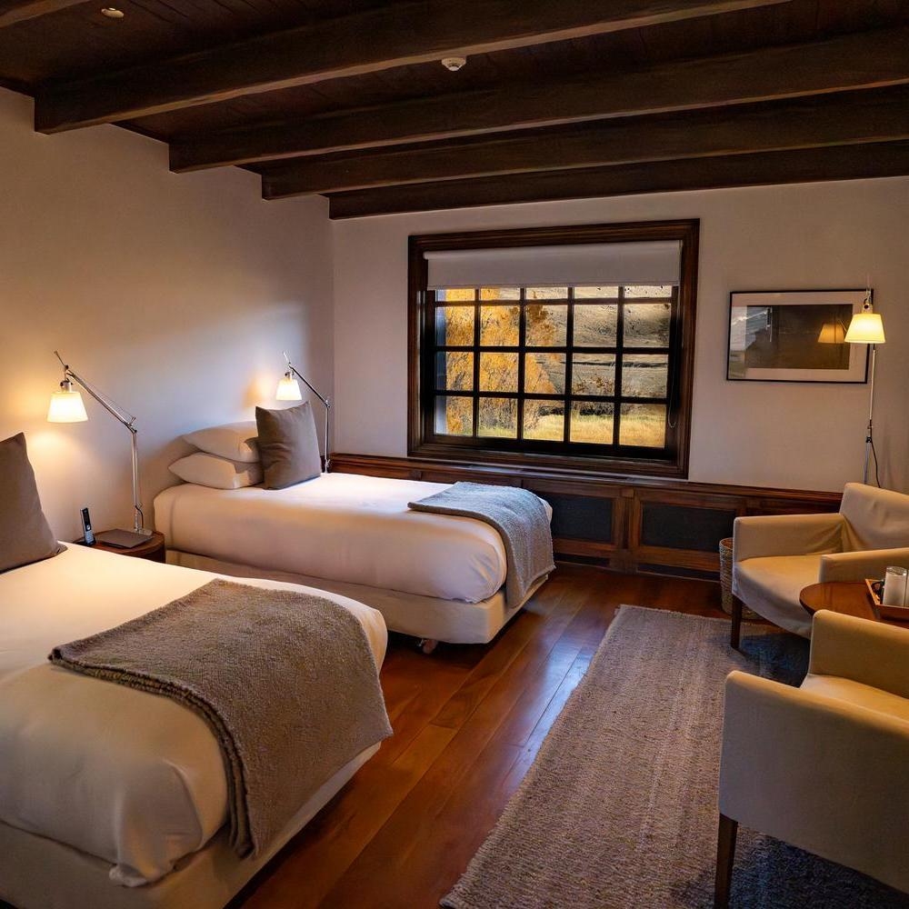 A twin bedroom with wooden ceiling beams, two beds, and armchairs next to a large grid window at Explora Patagonia National Park.