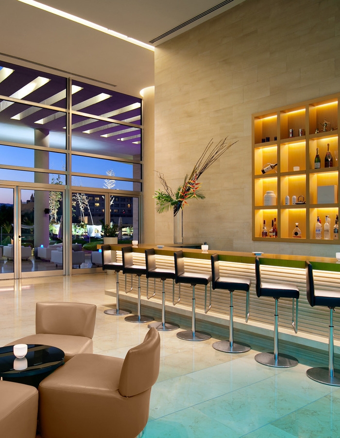Modern bar area at Kempinski Hotel Aqaba featuring black stools, lit shelves with bottles, and glass walls facing outdoors.