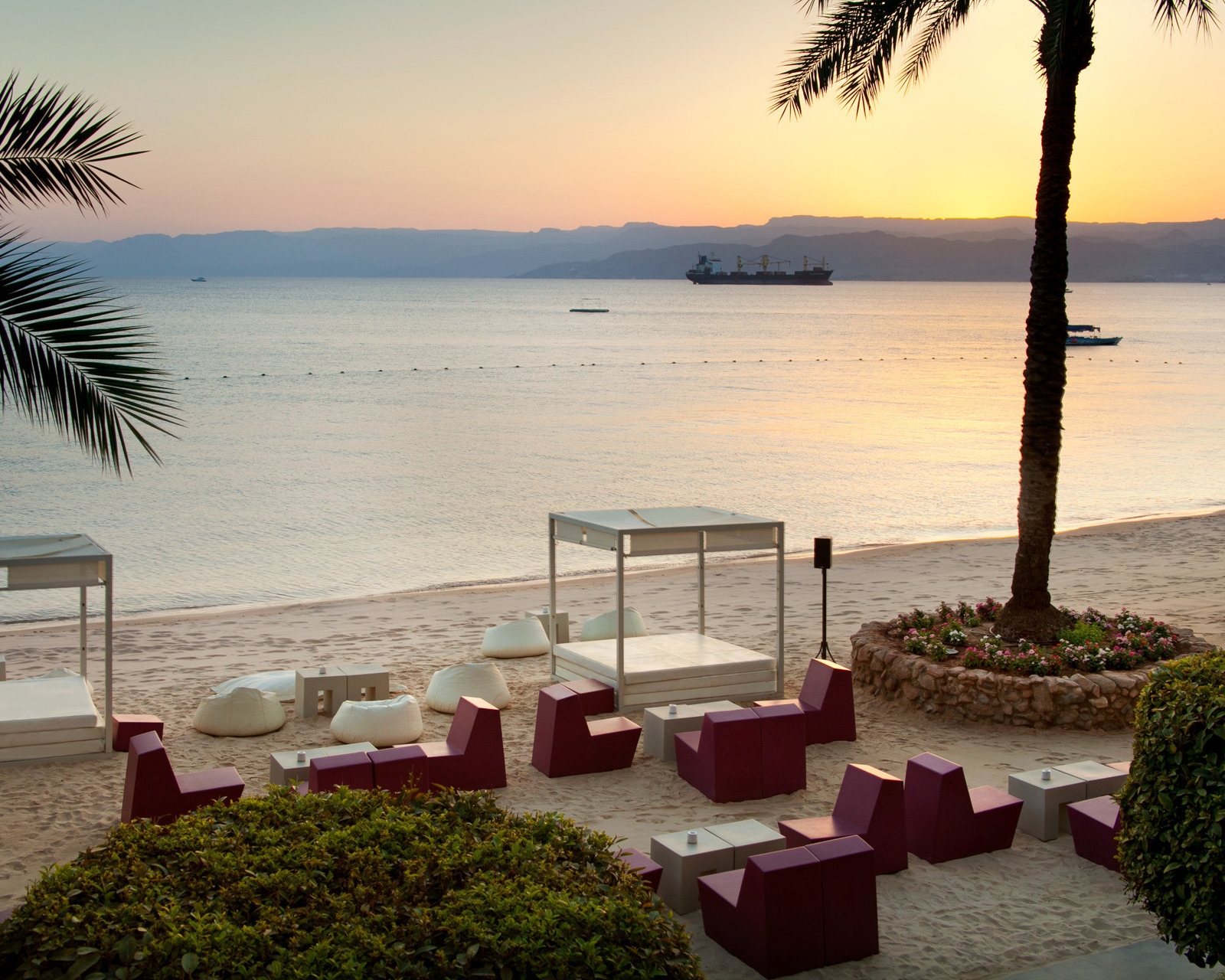 Sandy beach at Kempinski Hotel Aqaba featuring red block seats, white beanbags, and cabanas facing the sea at sunset.