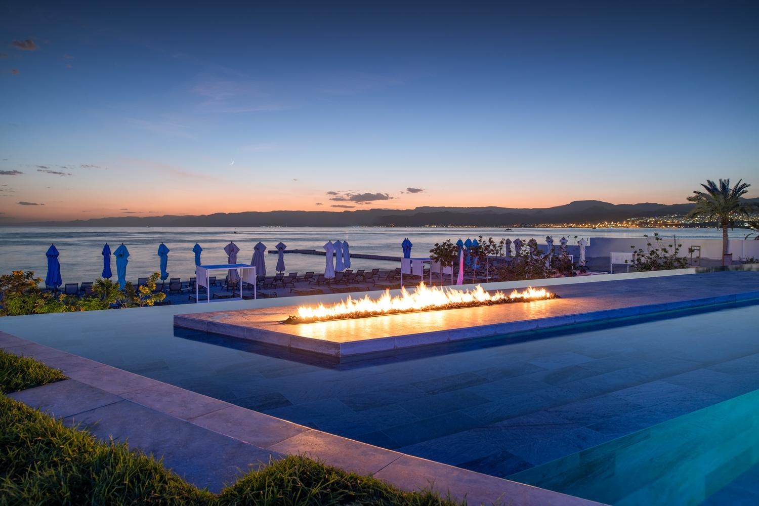 A long fire pit burning inside a modern infinity pool at twilight at Kempinski Hotel Aqaba, with sea views.