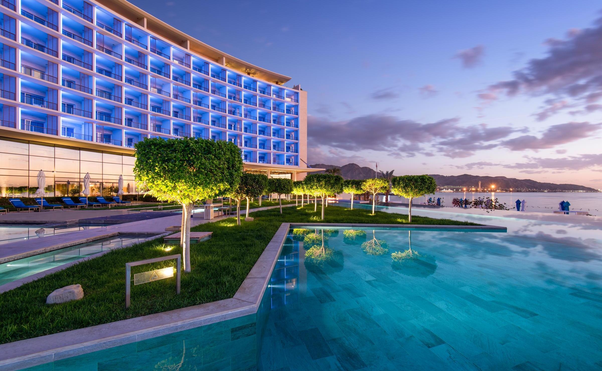 Curved exterior of Kempinski Hotel Aqaba glowing blue at dusk, reflecting in a large outdoor pool surrounded by trees.