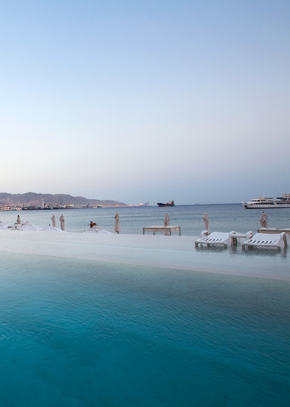 Calm infinity pool at dusk at Kempinski Hotel Aqaba with empty white lounge chairs resting in the shallow water.