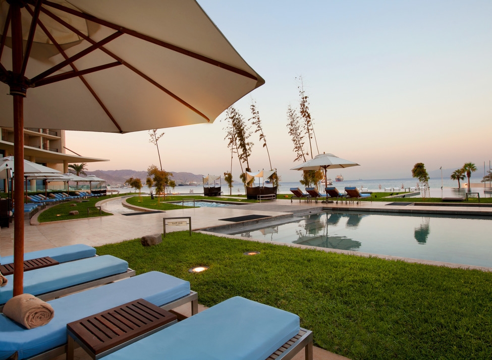 Blue lounge chairs and an umbrella by an outdoor pool at Kempinski Hotel Aqaba, overlooking the sea.
