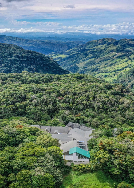 High-angle view of a large grey-roofed lodge surrounded by dense tropical forest and distant mountains.