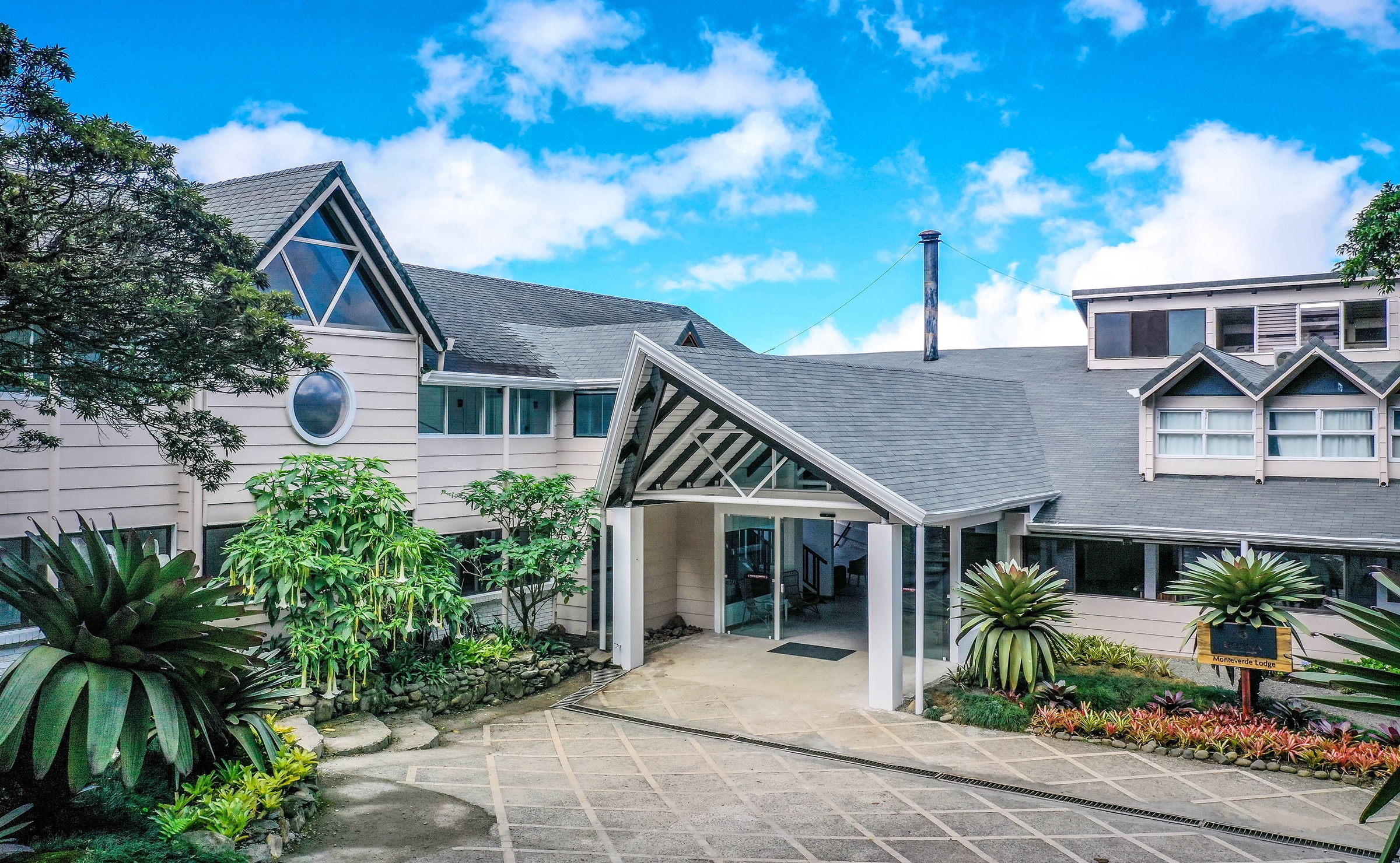 Exterior of Monteverde Lodge showing the white gabled entrance, stone pathways, and tropical plants.