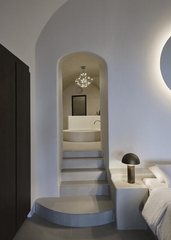 Archway with steps leading to a round soaking tub under a chandelier in a white bedroom at Andronis luxury suites.