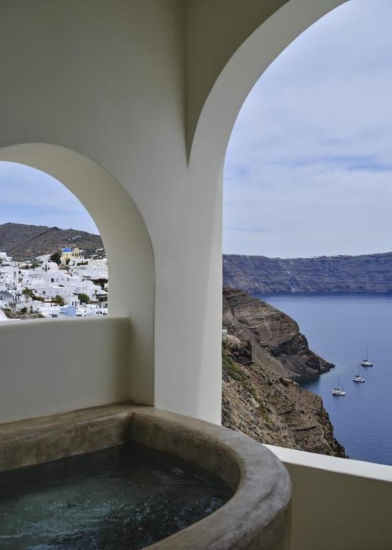View through arched windows at Andronis luxury suites showing a plunge tub, white buildings, and sea cliffs with boats.