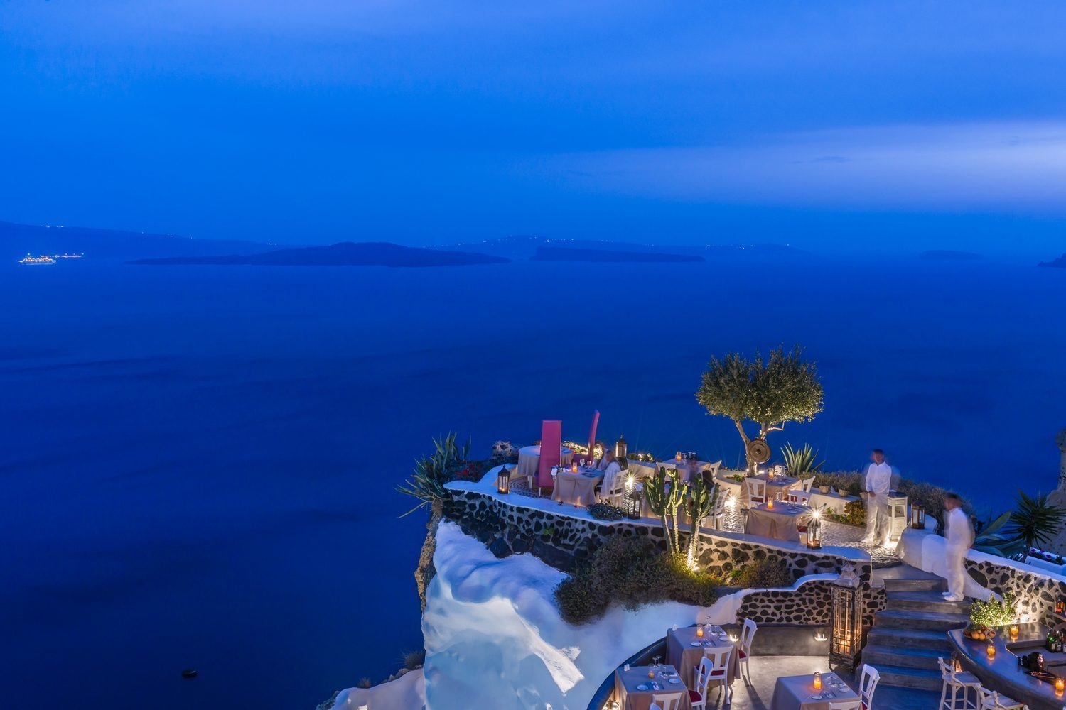 Twilight view of an outdoor cliffside restaurant at Andronis luxury suites with glowing lanterns, set tables, and sea views.