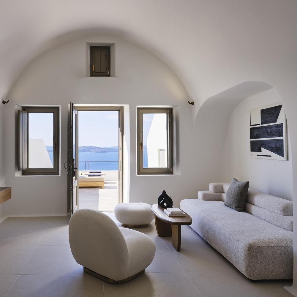 Bright living room at Andronis luxury suites with a grey sofa, white chairs, and open doors showing a sea-view balcony.