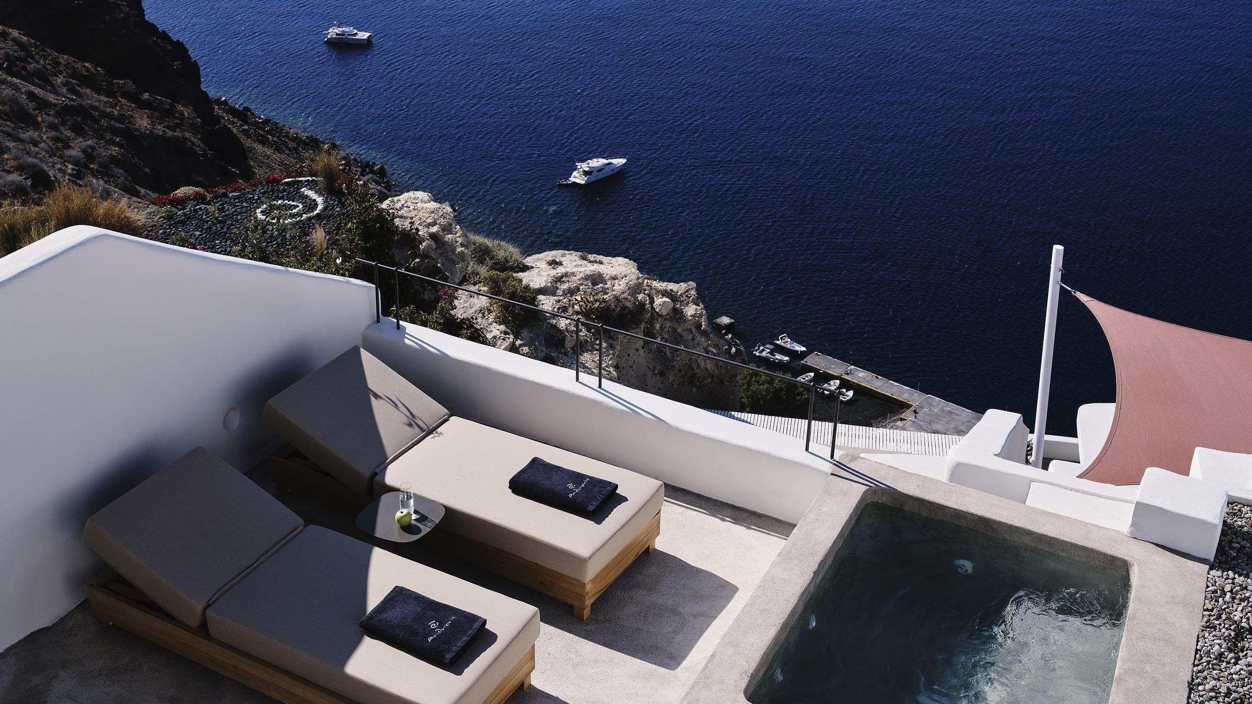 Elevated view of a balcony at Andronis luxury suites with two loungers, a plunge pool, and a view of the sea and cliffs.
