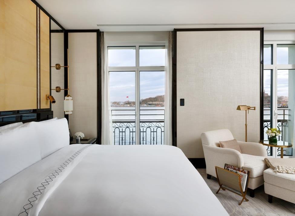 A modern white hotel bedroom with an armchair and glass doors opening to a view of the water and a distant flag.