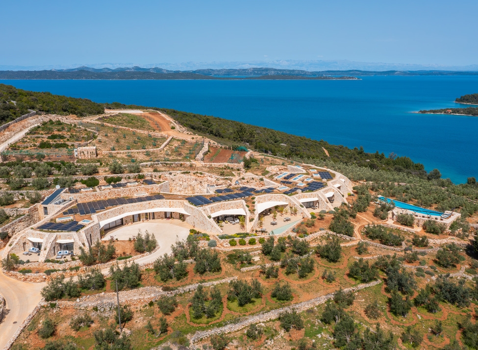 Panoramic aerial view of Villa Nai 3.3 built into a green coastal hillside under a clear blue sky.