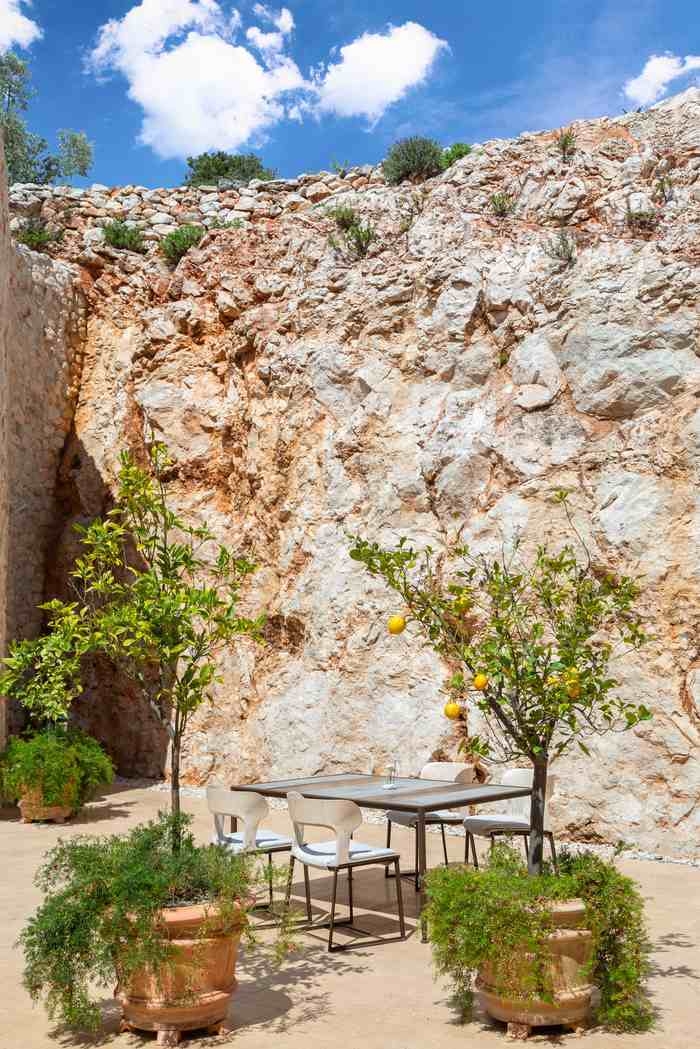 A modern dining table and chairs on a stone terrace in front of a tall, dramatic rock wall.