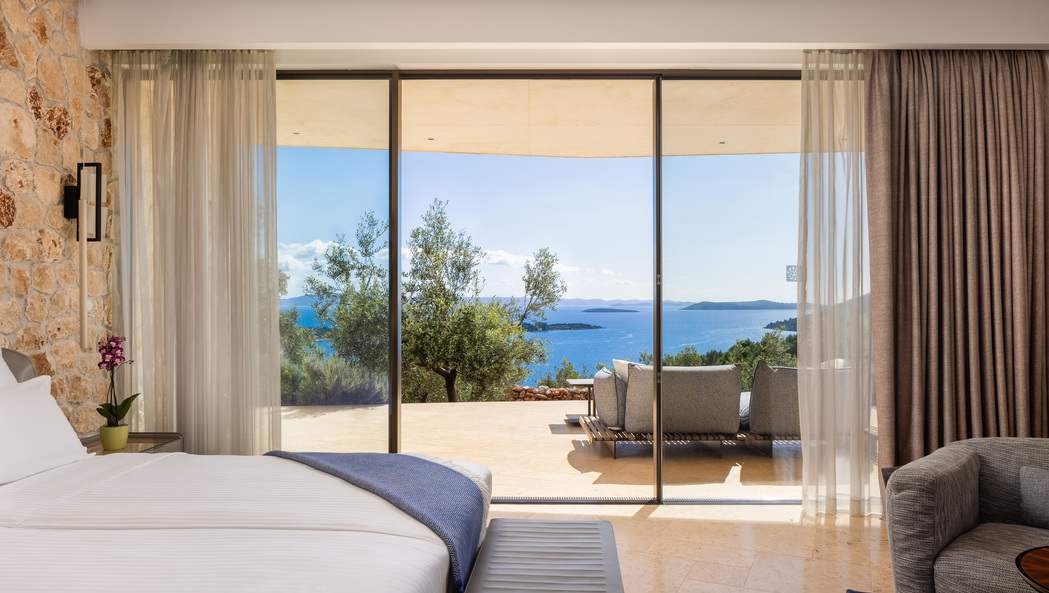 A modern bedroom with a large bed and glass doors opening to a terrace with panoramic sea views.