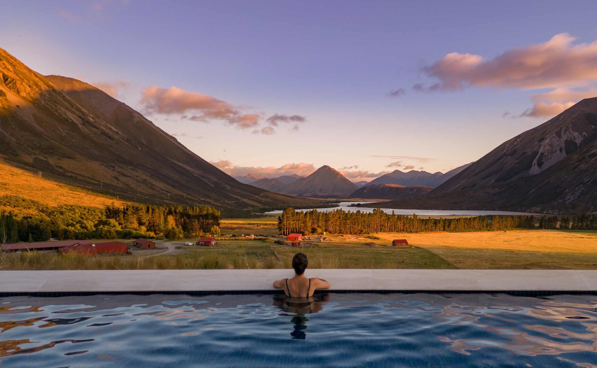 View from behind a person in an infinity pool looking out at a mountain valley landscape at dawn.