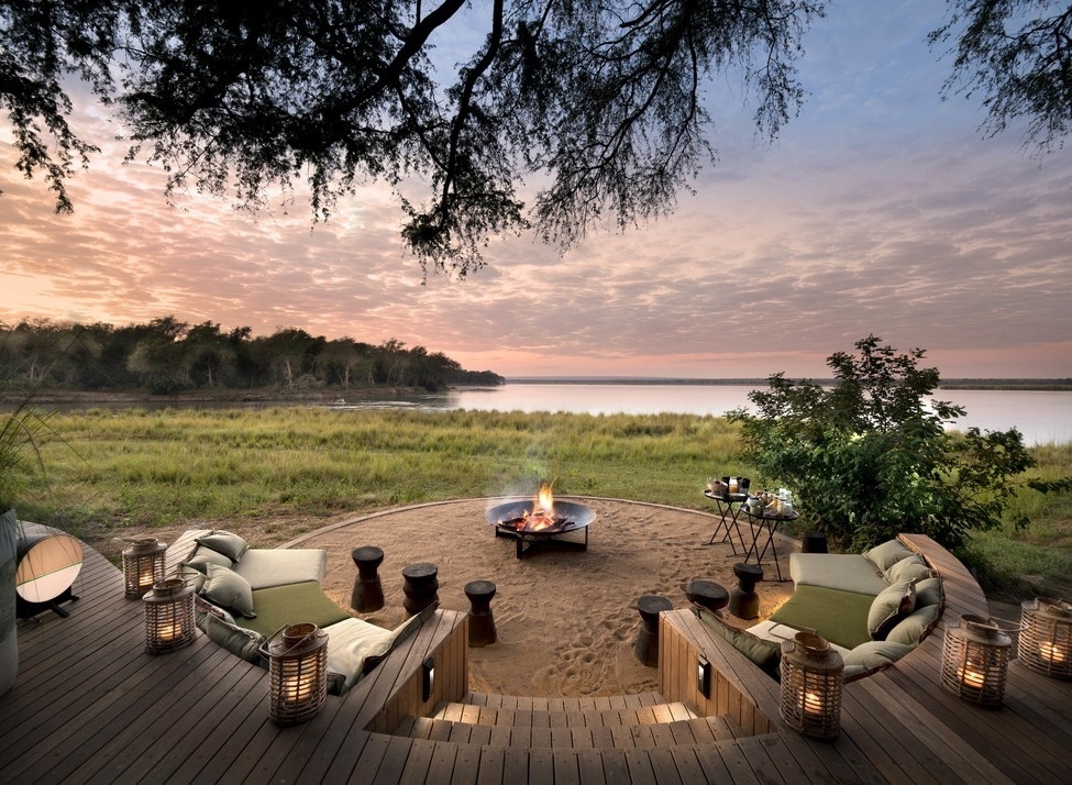 Sunken fire pit with curved seating on a wooden deck overlooking a grassy riverbank and calm river at sunset.