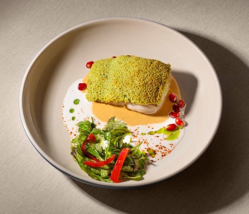 A high-angle shot of a plated fish dish with green crust, orange sauce, pomegranate, and greens.