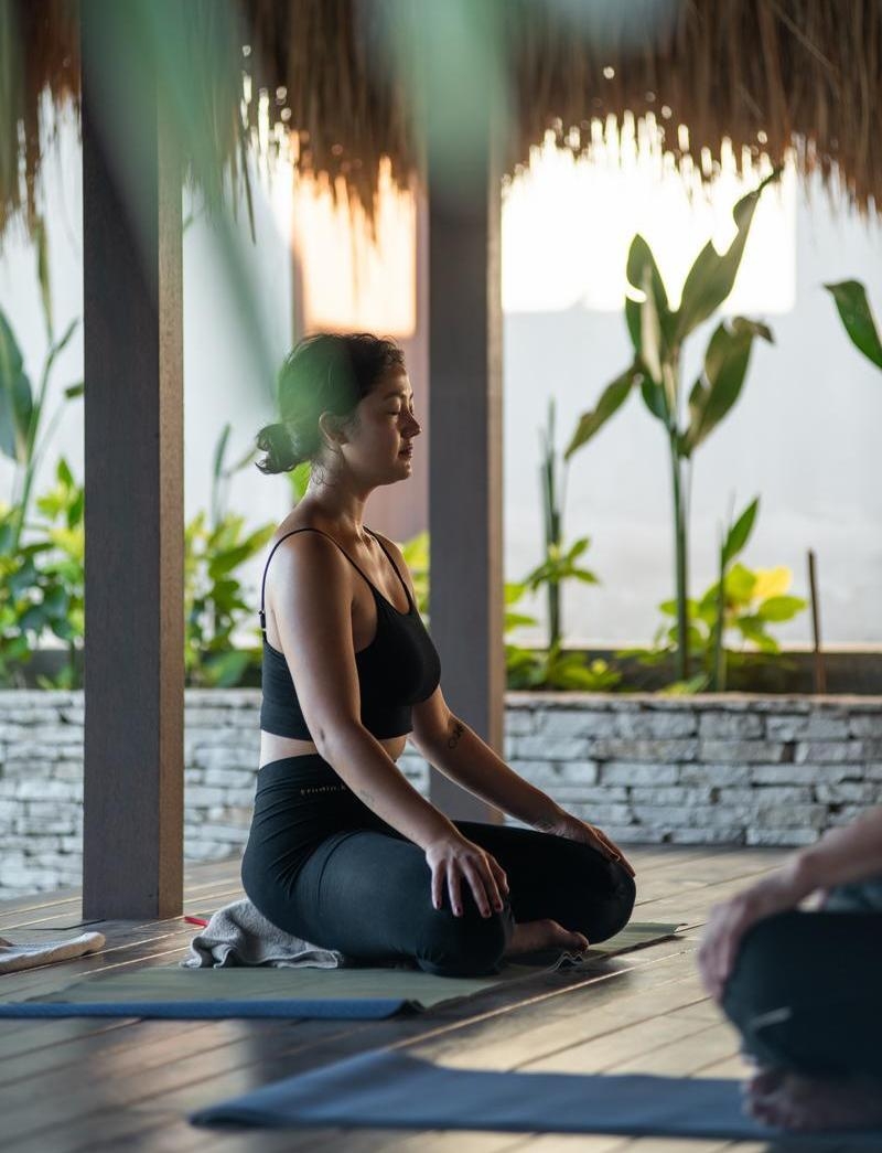 A woman practicing seated meditation on a yoga mat in a shaded, open-air tropical studio.