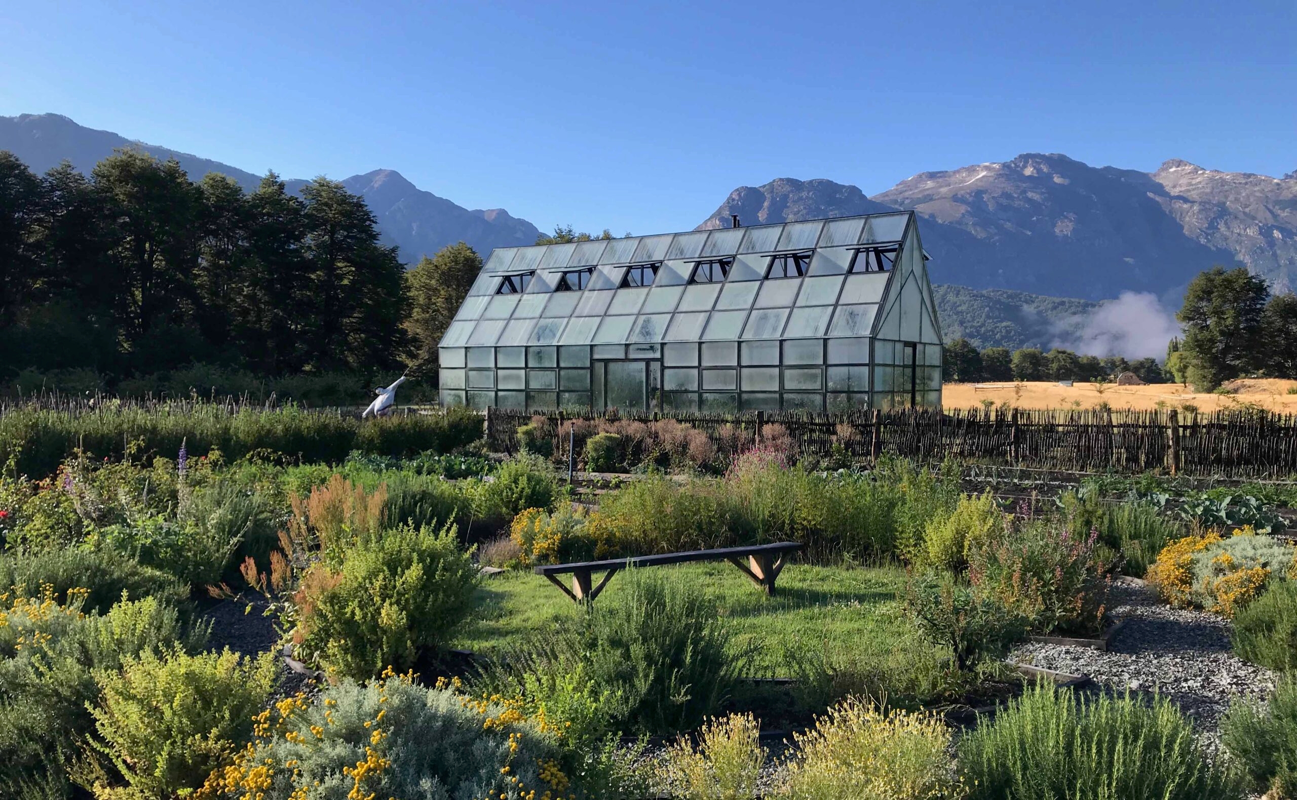 A greenhouse surrounded by lush gardens with mountains in the background under a clear sky.