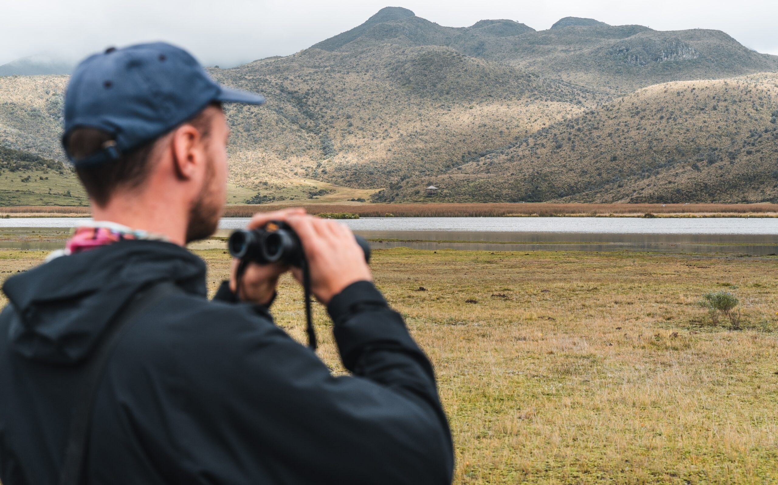Man with binoculars looking at a mountainous landscape across a lake.
