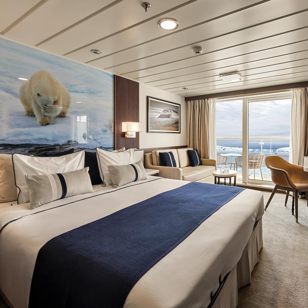 Hotel-style ship room with a large bed, seating area, and sliding glass doors opening to a balcony with icy ocean views.