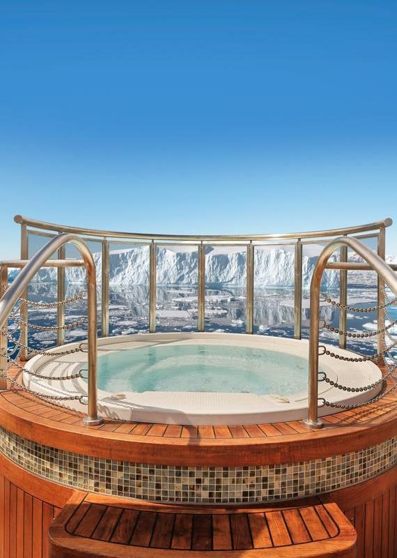 Round hot tub on a wooden ship deck enclosed by glass railings, looking out at a massive iceberg in calm, icy waters.