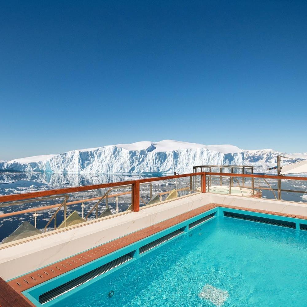Bright blue rectangular swimming pool on a ship deck, overlooking a massive tabular iceberg floating in calm icy waters.