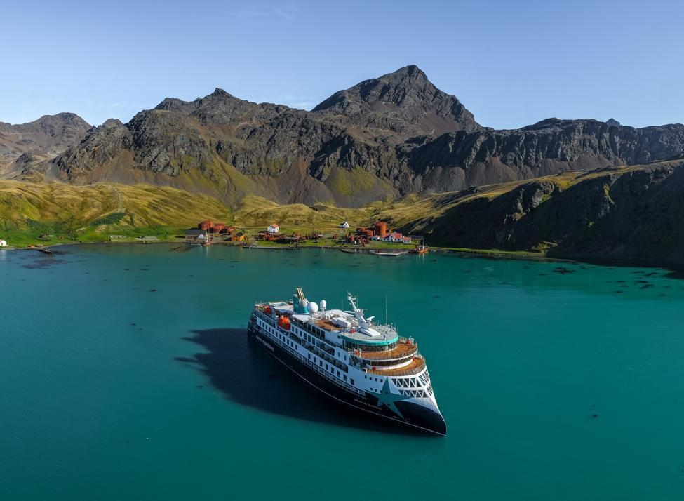 Large expedition cruise ship anchored in turquoise water near a shoreline with rusted buildings and rugged green mountains.