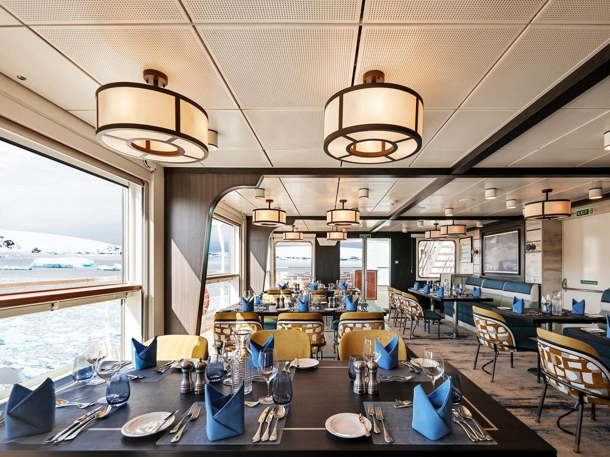 Bright dining room with set tables, blue napkins, and large windows looking out at an icy seascape.