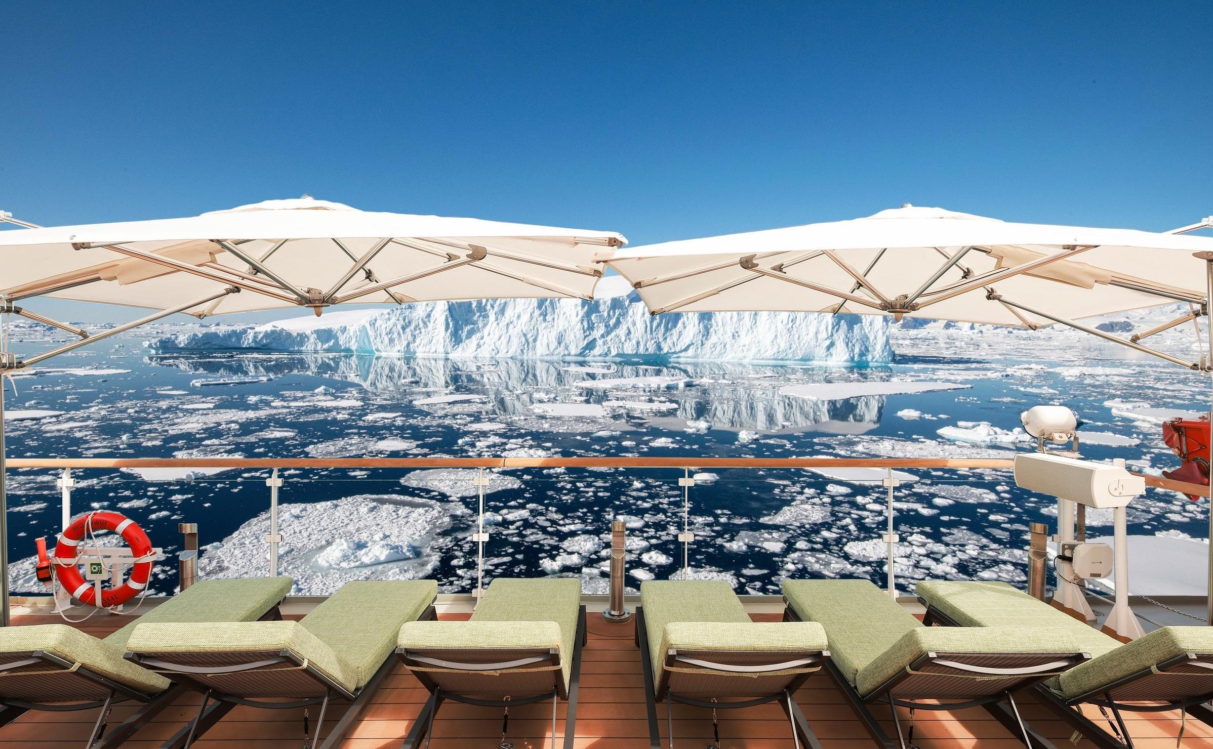 Ship deck with green lounge chairs, white umbrellas, and a life ring, facing a large iceberg surrounded by sea ice.