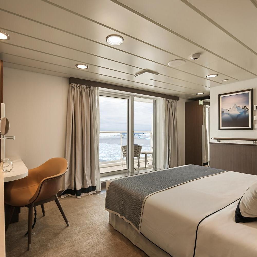 Ship cabin featuring a bed, desk with a brown chair, and sliding doors leading out to a balcony overlooking an icy sea.