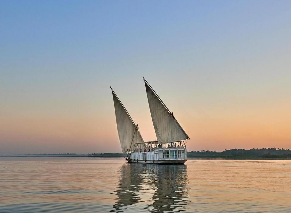 A traditional white twin-masted Dahabiya boat sailing on the calm Nile River at sunset with a clear orange and blue sky.