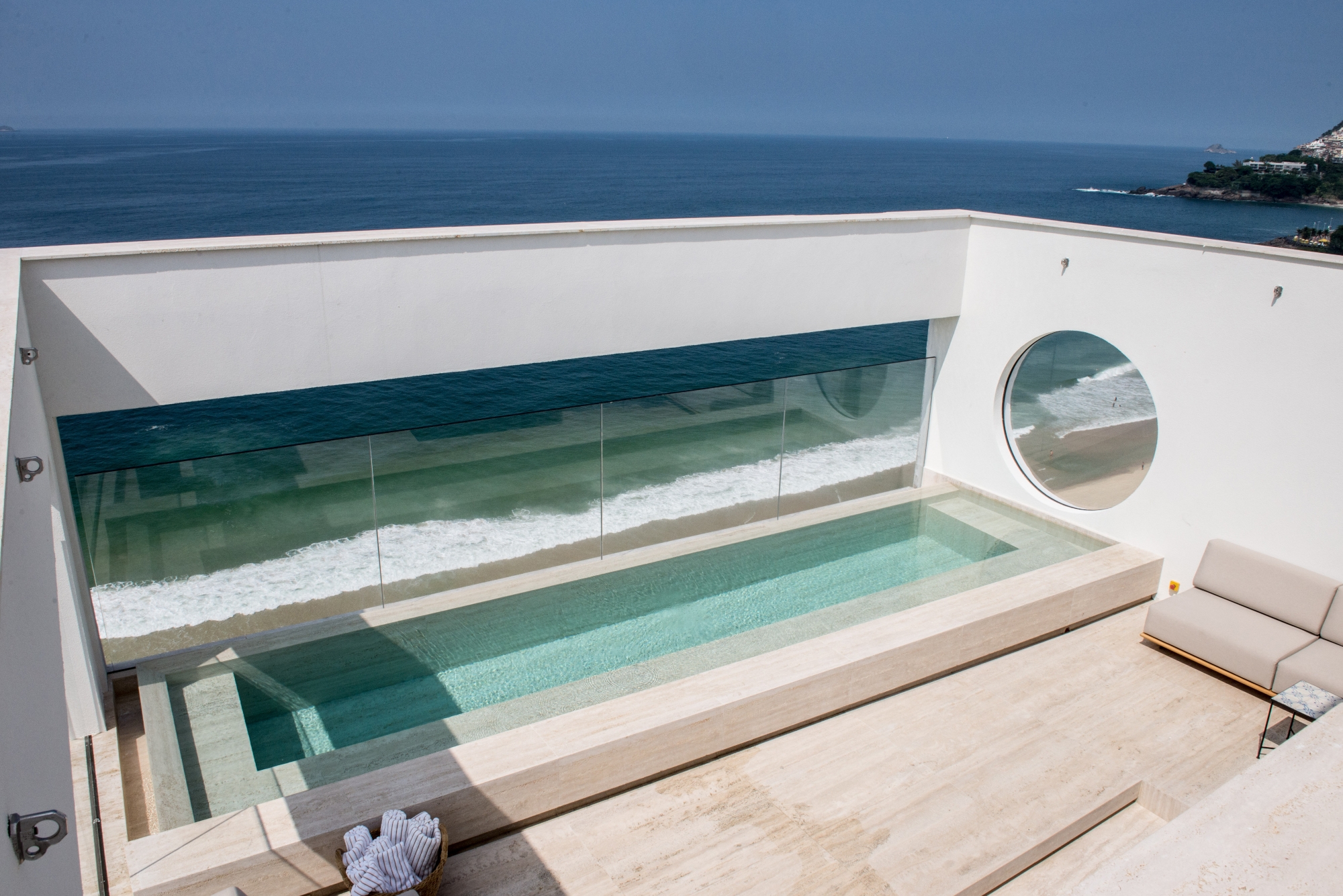 View of a pool and beige lounge area from above. The pool has windows overlooking a beach and seafront.