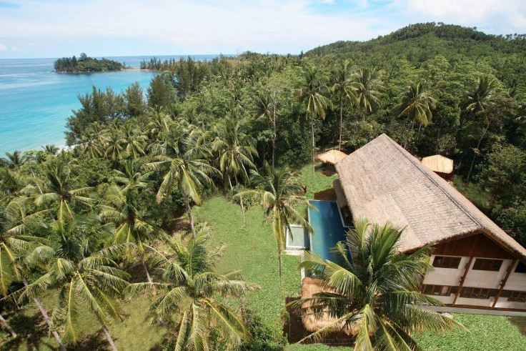 Aerial shot of a villa with thatched roof surrounded by a small lawn and a palm forest, with a blue sea in the background.