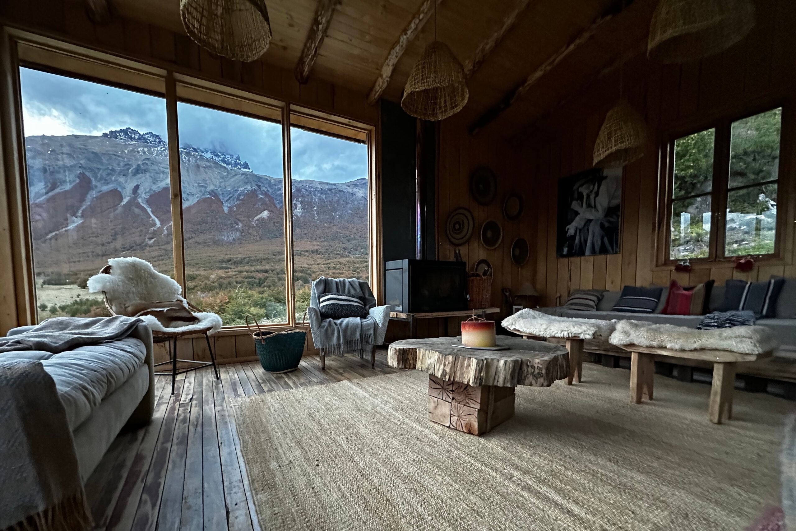 Spacious living room with large picture windows facing snow-capped mountains, woven pendant lamps, cushioned seating and a low table made from a tree slab.