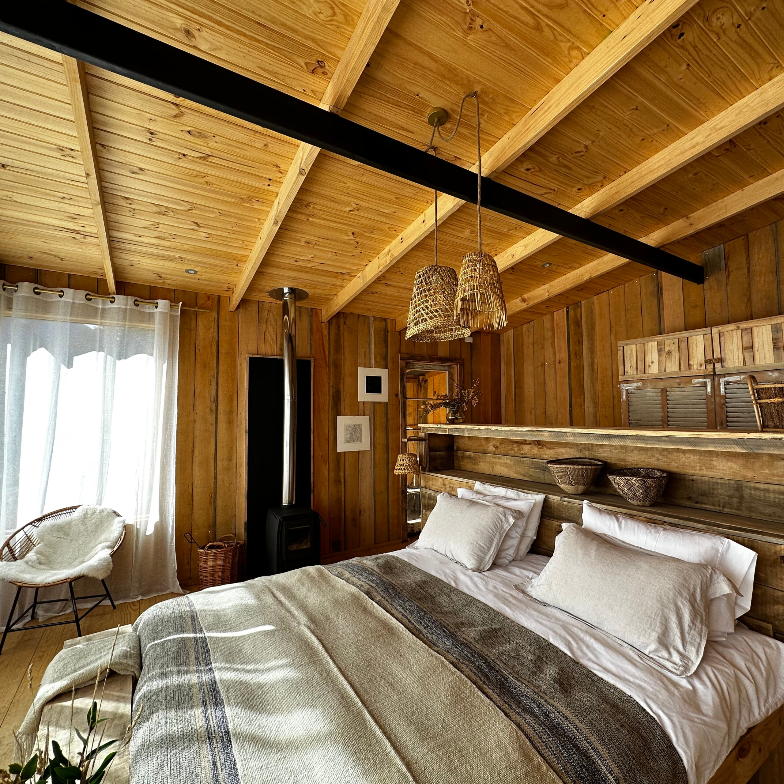 Bedroom with a wooden ceiling and walls, a large bed with neutral linens, woven pendant lights, a wood burner and sheer curtains filtering natural light.