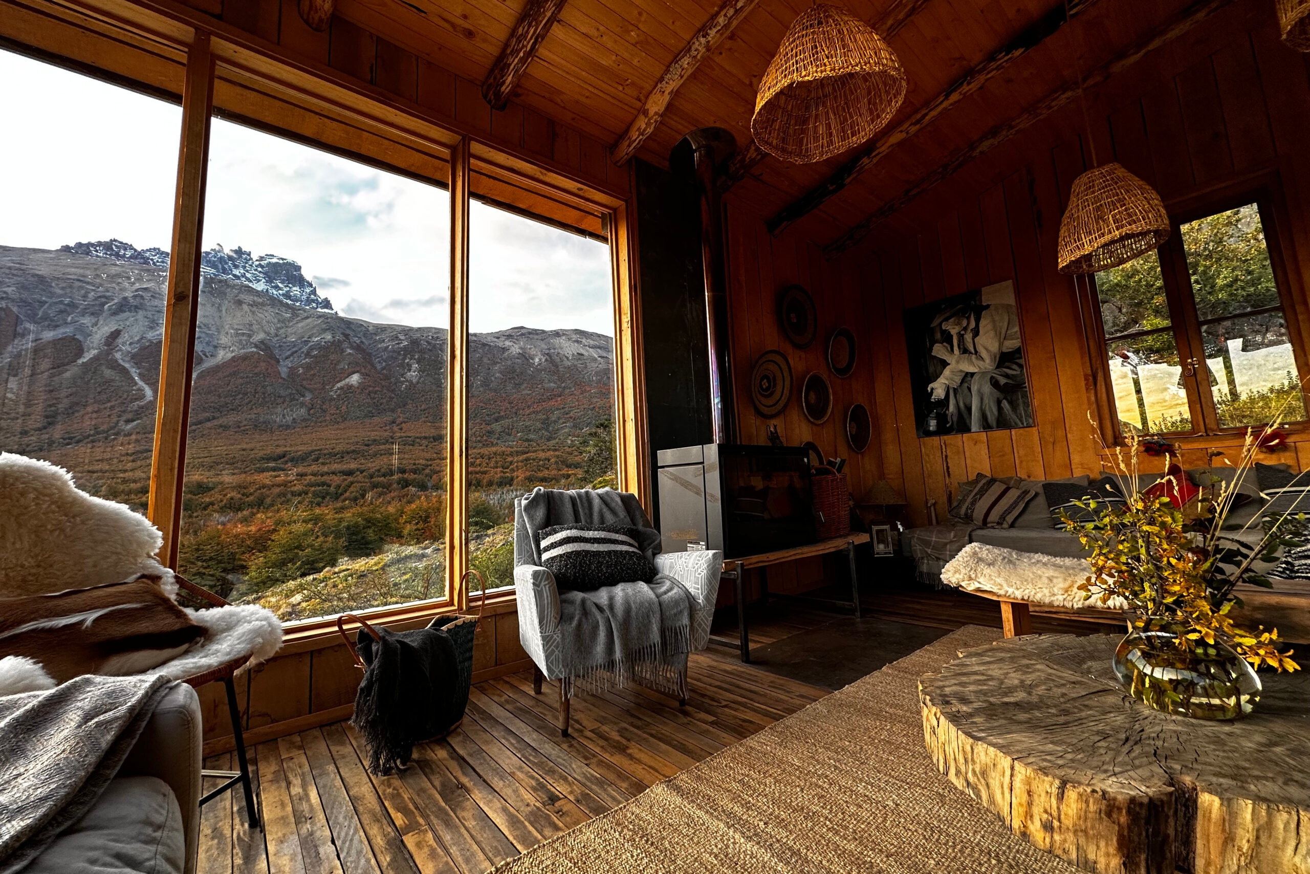 Lounge area with wooden floors, large windows framing a mountain slope, cushioned armchairs with throws, and a wood stove set against timber walls.