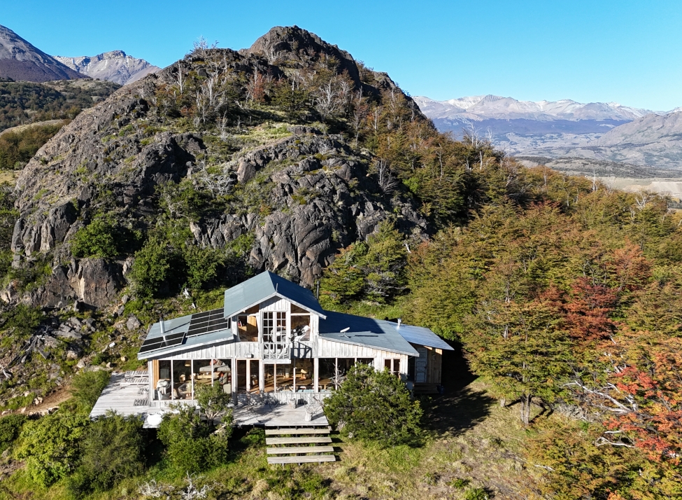 Two-storey cabin with pitched grey roof and steps leading up to the front sits in front of a large grey rock formation on a green mountain slope with mountains in the background.