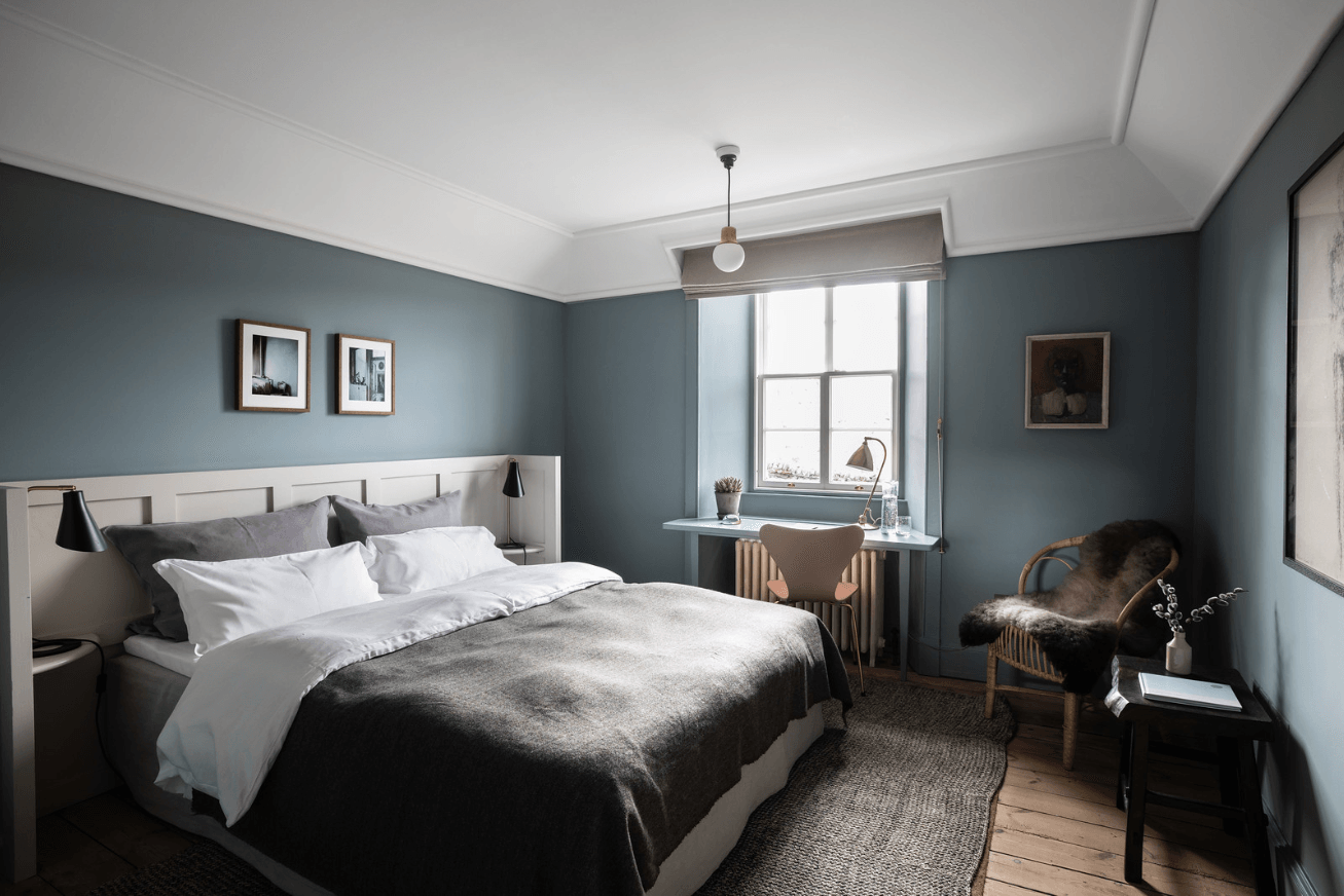 A guest room with grey-blue walls, a large bed, a desk facing a window and a chair with a fur blanket on it