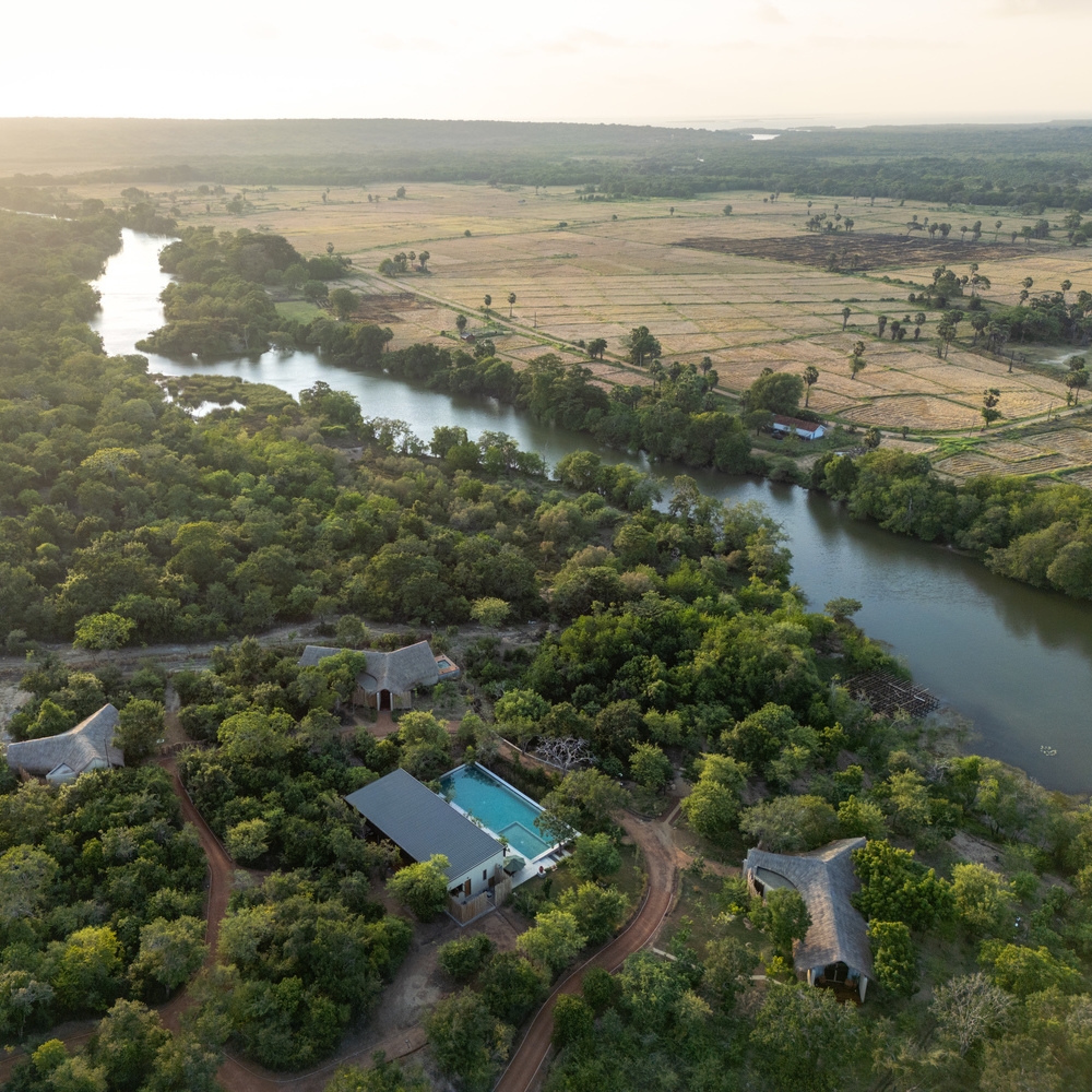 Drone shot of a river winding through a forest with several thatched-roof buildings and a swimming pool.