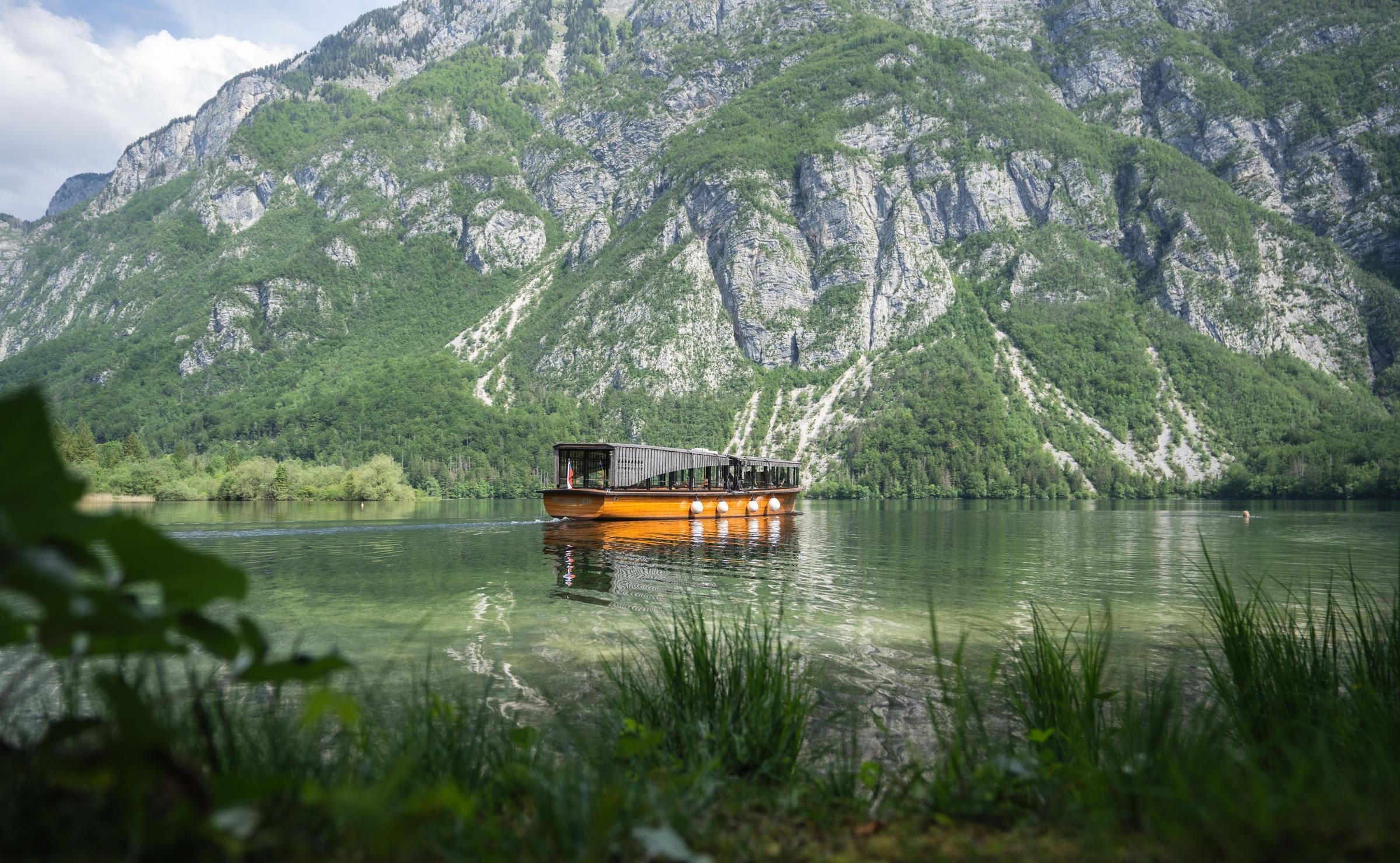 A vintage wooden tourist boat on a calm green lake surrounded by steep, forested mountains under a bright sky.