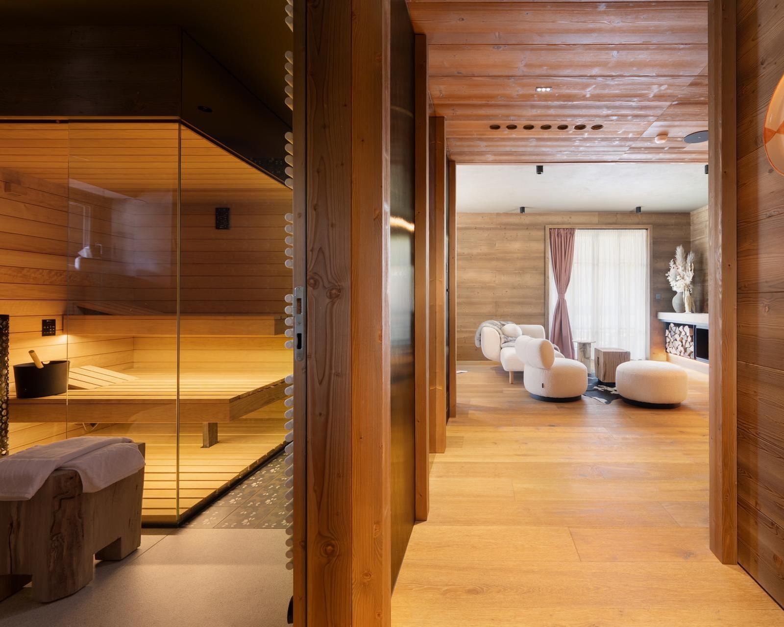 Modern hotel suite interior showing a glass sauna on the left and a bright living room with white chairs.
