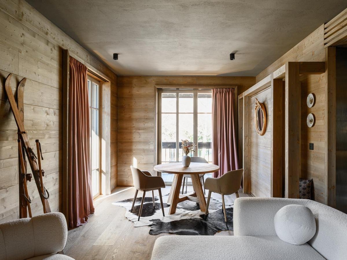 Sunlit room with wood walls, a round dining table on a cowhide rug, and vintage skis leaning against the wall.